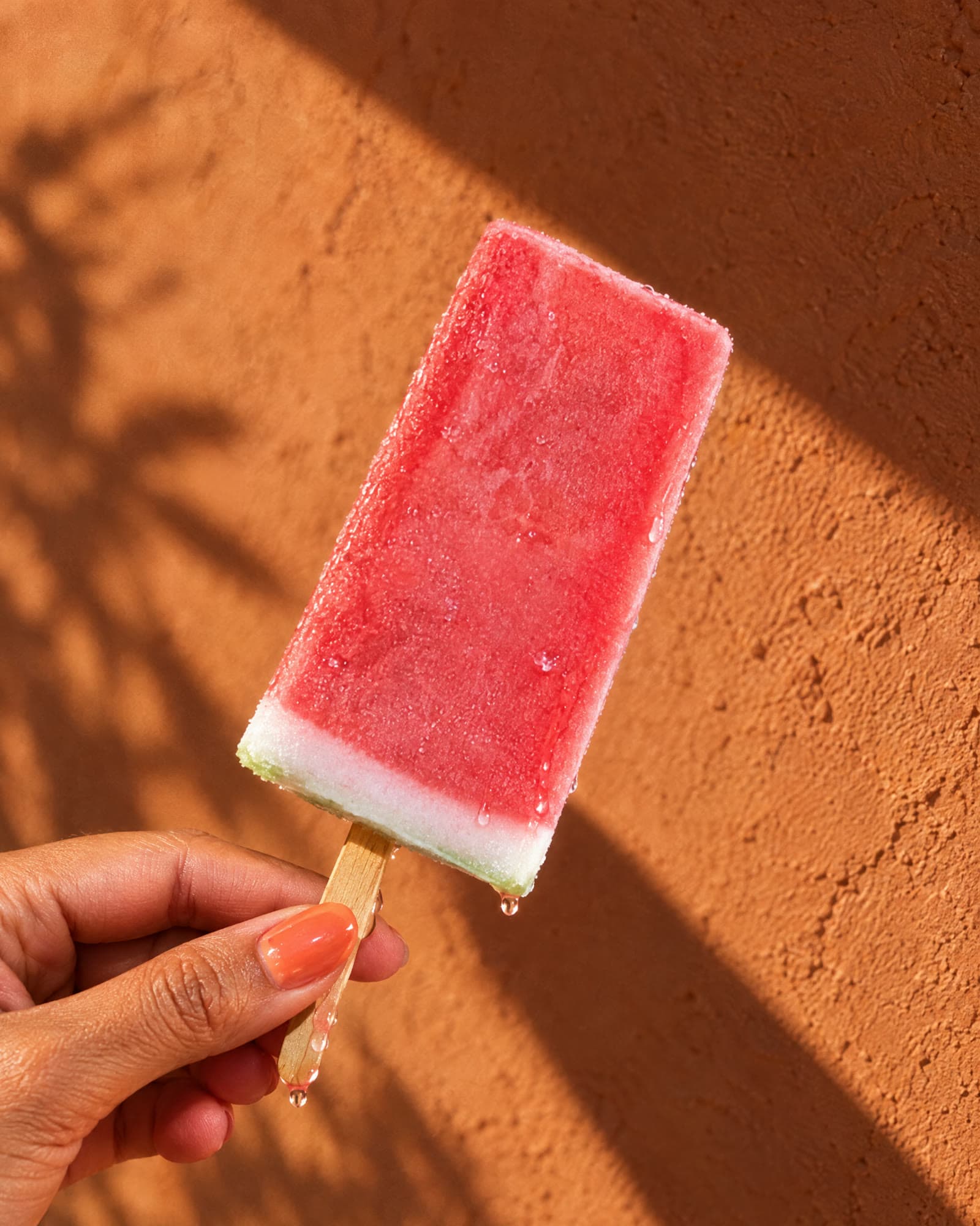 A hand holding a watermelon popsicle against a sunlit terracotta wall.